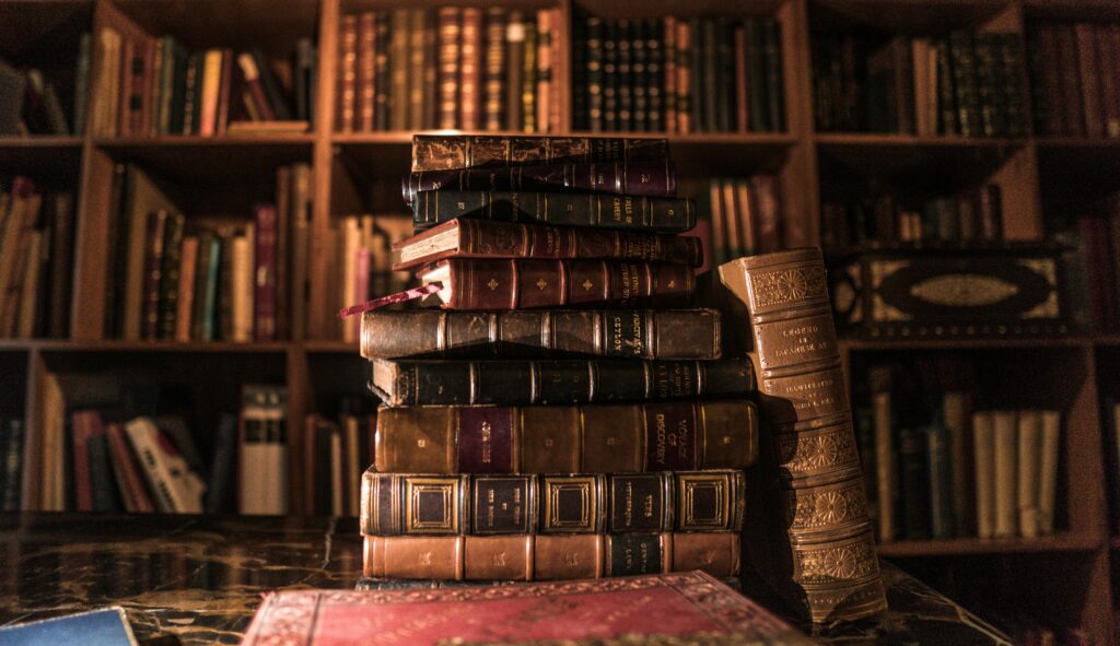 A pile of leather bound books in front of a full set of wooden shelves, symbolizing the research behind Morgan's A. Drake non-fiction maritime myths and legends, and literary critique works, featured on fathoms deep on substack.